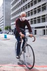 Hombre caucásico mayor por las calles de la ciudad durante el día, con una máscara facial contra el coronavirus, covid 19, montando su bicicleta. - foto de stock