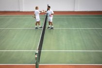 Un Caucasien et un homme de race mixte portant des blancs de tennis passant du temps sur un court ensemble, jouant au tennis par une journée ensoleillée, serrant la main, tenant une raquette de tennis — Photo de stock