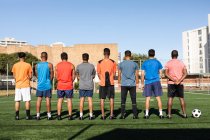 Vista trasera de varios grupos étnicos de hombres cinco jugadores de fútbol de lado con entrenamiento de ropa deportiva en un campo de deportes bajo el sol, de pie en una fila antes de un juego con pelota junto a ellos. - foto de stock