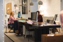 Creativos de negocios caucásicos femeninos y masculinos que trabajan en una oficina moderna informal, sentados en escritorios y usando computadoras, tomando notas - foto de stock