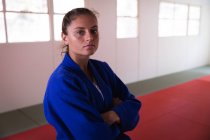 Portrait of a confident teenage Caucasian female judoka wearing blue judogi, standing in the gym with her arms crossed and looking straight into a camera. — Stock Photo