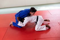 Vue latérale d'un entraîneur de judo masculin de race mixte et d'un judoka masculin de race mixte adolescent portant du judogi bleu et blanc, pratiquant le judo lors d'un entraînement dans une salle de gym. — Photo de stock