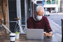 Hombre caucásico mayor sentado en una mesa en una terraza de café, con una máscara facial contra coronavirus, covid 19, usando un teléfono inteligente y un ordenador portátil. - foto de stock