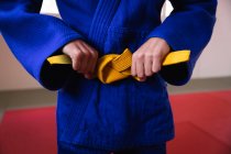 Front view mid section of judoka standing on gym mats, tying up the yellow belt of blue judogi. — Stock Photo