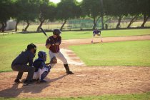 Вид сзади на белого бейсболиста во время игры в бейсбол в солнечный день, готовящегося ударить по мячу бейсбольной битой, кэтчер и другой игрок сидят на корточках за киллером — стоковое фото