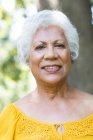 Портрет крупным планом привлекательной пожилой афроамериканки с короткими белыми волосами, наслаждающейся уходом на пенсию в саду на солнце, смотрящей в камеру и улыбающейся, самоизолирующейся во время пандемии коронавируса — стоковое фото