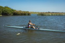 Side view of a Caucasian male rower training and rowing on the river, holding oars and sitting in a rowing boat on a sunny day — Stock Photo