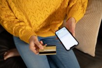 Hochwinkel-Mittelteil einer Frau, die es sich zu Hause gemütlich macht, auf einem Sofa sitzt, eine Kreditkarte in der Hand hält und mit ihrem Smartphone eine Online-Transaktion tätigt — Stockfoto