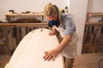 Caucasian male surfboard maker with long blond hair, wearing a face mask, working in his studio, making a surfboard, inspecting it and preparing to polishing.. — Stock Photo
