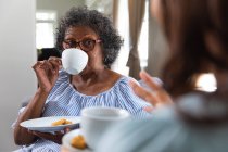 Mujer de raza mixta mayor pasando tiempo en casa con su hija, distanciamiento social y aislamiento en cuarentena, tomando el té juntos y hablando - foto de stock
