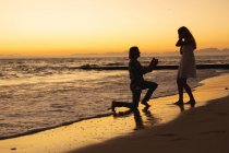 Coppia caucasica godendo il loro tempo in una spiaggia durante il tramonto, un uomo si inginocchia e propone a una donna — Foto stock