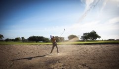 Vista lateral de um homem caucasiano em um campo de golfe em um dia ensolarado com céu azul, batendo uma bola de golfe — Fotografia de Stock
