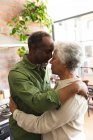 Вид сбоку счастливой пожилой афроамериканской пары в отставке, стоящей дома на кухне, трогающей головы и смотрящей друг на друга, обнимаясь, дома вместе изолирующей во время пандемии коронавируса — стоковое фото