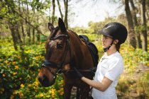 Vista laterale da vicino di una cavallerizza caucasica vestita con disinvoltura che conduce un cavallo di castagno lungo un sentiero attraverso una foresta durante una giornata di sole. — Foto stock