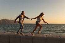 Casal caucasiano em umas férias de praia andando em um passeio marítimo durante o pôr do sol, de mãos dadas, com o céu e o mar no fundo — Fotografia de Stock