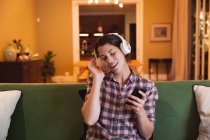 Mujer de raza mixta pasar tiempo en casa auto aislamiento y distanciamiento social en cuarentena de bloqueo durante coronavirus covid 19 epidemia, el uso de auriculares celebración de teléfono inteligente en la sala de estar. - foto de stock