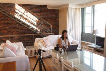Vlogger femminile caucasica a casa, nel suo salotto utilizzando un computer portatile per preparare il suo blog online. Distanziamento sociale e autoisolamento in quarantena. — Foto stock
