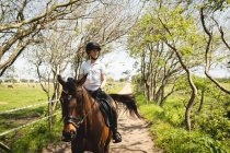 Vista frontale di una cavallerizza caucasica vestita con disinvoltura che hackera un cavallo di castagno su un sentiero attraverso una foresta in una giornata di sole. — Foto stock