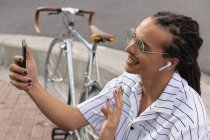 Seitliche Nahaufnahme eines gemischten Rennfahrers mit langen Dreadlocks, der an einem sonnigen Tag in der Stadt unterwegs ist, mit Kopfhörern auf der Straße sitzt, sein Smartphone benutzt und mit der Hand wedelt, während sein Fahrrad neben ihm steht.. — Stockfoto