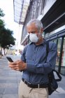 Homem caucasiano sênior nas ruas da cidade durante o dia, usando uma máscara facial contra coronavírus, vívido 19 e usando um smartphone . — Fotografia de Stock