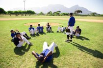 Vista lateral de um grupo multi-étnico de jogadores de beisebol do sexo masculino segurando os pés uns dos outros, treinando antes de um jogo, exercitando-se fazendo sit ups em um círculo, em um campo de jogo em um dia ensolarado — Fotografia de Stock