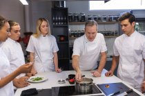 Grupo caucasiano de chefs masculinos e femininos, ouvindo um chef caucasiano sênior adicionando ingredientes a um pote. Aula de culinária em uma cozinha de restaurante. — Fotografia de Stock