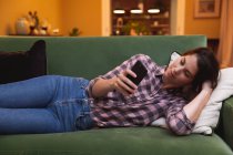 Mixed race woman spending time at home self isolating and social distancing in quarantine lockdown during coronavirus covid 19 epidemic, lying on a sofa using smartphone in sitting room. — Stock Photo