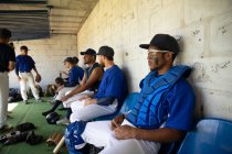 Vista lateral de una fila de jugadores de béisbol masculinos multiétnicos, preparándose antes de un juego, sentados en el vestuario, enfocándose mientras esperan, interactuando - foto de stock
