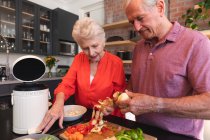 Счастливая пожилая кавказская пара дома, готовит еду и улыбается на кухне, мужчина режет овощи, женщина смотрит и разговаривает с ним, дома вместе изолируя во время пандемии коронавируса — стоковое фото