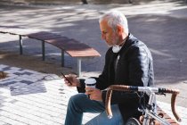 Homme caucasien âgé dehors et autour dans les rues de la ville pendant la journée, portant un masque facial contre le coronavirus, covid 19, assis sur un banc, tenant une tasse de café à emporter et en utilisant son smartphone. — Photo de stock