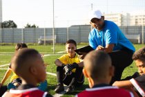 Frontansicht eines gemischten männlichen Fußballtrainers, der während eines Fußballtrainings auf einem Spielfeld in der Sonne kniet und eine multiethnische Gruppe von Jungen-Fußballern instruiert — Stockfoto