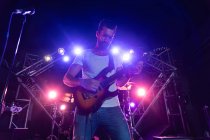 Фронт-вью крупным планом кавказского гитариста, сосредоточенного на игре на электрогитаре, стоящего на прожекторной сцене с группой, выступающей на музыкальной площадке, с бас-гитаристом и ударной установкой на заднем плане — стоковое фото