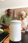 Счастливая пожилая афроамериканская пара дома, готовит овощи для еды, контейнер для компостирования зеленых отходов на переднем плане, пара дома вместе изолирует во время пандемии коронавируса — стоковое фото