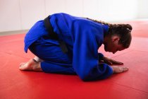 Vue latérale d'une judoka blanche adolescente portant du judogi bleu, agenouillée et s'inclinant sur des nattes dans la salle de gym avant l'entraînement du judo. — Photo de stock