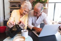 Una pareja de ancianos jubilados caucásicos en casa de pie en una mesa en su cocina, hablando y sonriendo, utilizando un ordenador portátil juntos y sonriendo, la mujer vertiendo café de una olla, pareja aislante durante coronavirus covidemic 19 - foto de stock