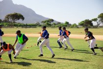 Seitenansicht einer multiethnischen Gruppe männlicher Baseballspieler, die an einem sonnigen Tag mit ihrem Trainer auf einem Spielfeld trainieren, kurze Sprints machen und sich drehen. — Stockfoto