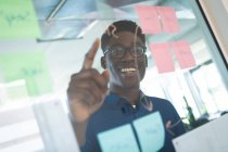 Un uomo d'affari afroamericano che indossa una camicia blu e occhiali, lavora in un ufficio moderno, scrive su lavagna trasparente con appunti e sorride — Foto stock