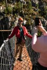 Sur la vue d'épaule du couple caucasien profitant du temps dans la nature, portant un équipement de tyrolienne, la femme prenant des photos avec un smartphone par une journée ensoleillée dans les montagnes — Photo de stock