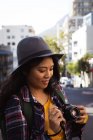 Side view close up of a mixed race woman with long dark hair out and about in the city streets during the day, using a digital camera, wearing a hat and checked shirt and walking in a city street with buildings in the background. — Stock Photo