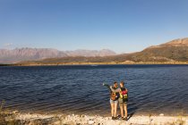 Visão traseira de um casal caucasiano se divertindo em uma viagem às montanhas, de pé em um caminho, em uma costa, uma mulher está apontando para algo, em um dia ensolarado. — Fotografia de Stock