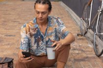 Vista frontal de cerca de un hombre de raza mixta con rastas largas fuera y alrededor de la ciudad en un día soleado, sentado en la calle, utilizando un teléfono inteligente y sosteniendo una taza de café con su bicicleta de pie junto a él. - foto de stock