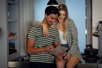 Side view of a young mixed race man and a young Caucasian woman sitting in the kitchen, embracing themselves, smiling and using their tablet. — Stock Photo
