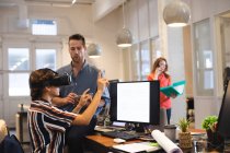 Смешанная раса женщин бизнес-креатив, работающих в обычном современном офисе, сидя за столом с помощью гарнитуры VR с коллегами, работающими на заднем плане — стоковое фото