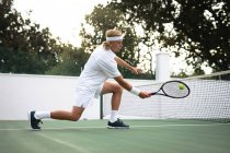 Un homme caucasien portant des blancs de tennis passe du temps sur un court à jouer au tennis par une journée ensoleillée, tenant une raquette de tennis, frappant une balle — Photo de stock