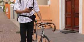Vue de face section médiane de l'homme avec de longs dreadlocks dehors et autour dans la ville par une journée ensoleillée, marcher dans la rue à l'aide d'un smartphone et rouler son vélo. — Photo de stock