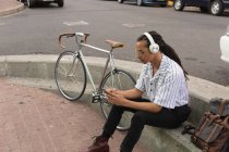 Вид сбоку на смешанного гонщика с длинными дредами в городе в солнечный день, сидящего на улице в наушниках и использующего смартфон с велосипедом, стоящим рядом с ним. — стоковое фото