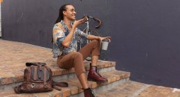 Vista lateral de un hombre de raza mixta con largas rastas en la ciudad en un día soleado, sentado en la calle y sonriendo, usando un teléfono inteligente y sosteniendo una taza de café, con su bicicleta apoyada en la pared junto a él. - foto de stock