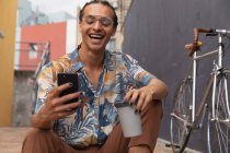 Вид спереди на смешанного гонщика с длинными дредами в солнечный день, сидящего на улице и улыбающегося, с помощью смартфона и с чашкой кофе, с велосипедом, стоящим рядом с ним. — стоковое фото