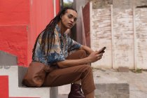 Side view of a mixed race man with long dreadlocks out and about in the city on a sunny day, sitting on the stairs in the street and using a smartphone, looking straight into camera. — Stock Photo