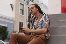Vista frontal de cerca de un hombre de raza mixta con rastas largas en la ciudad en un día soleado, sentado en las escaleras en la calle y usando un teléfono inteligente, mirando directamente a la cámara. - foto de stock